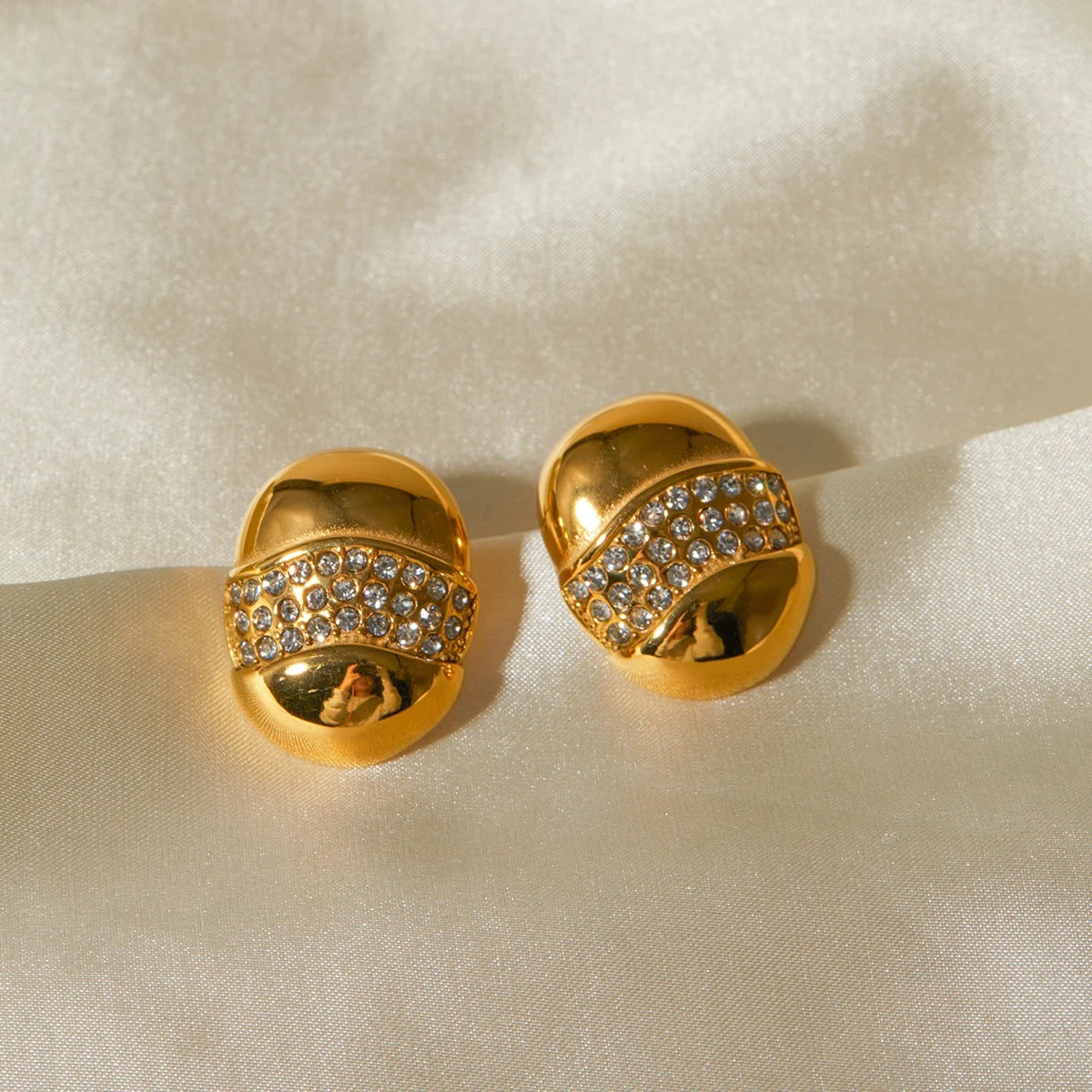 A pair of gold-tone dome-shaped earrings with a wave-like band encrusted with sparkling rhinestones on a white background.
