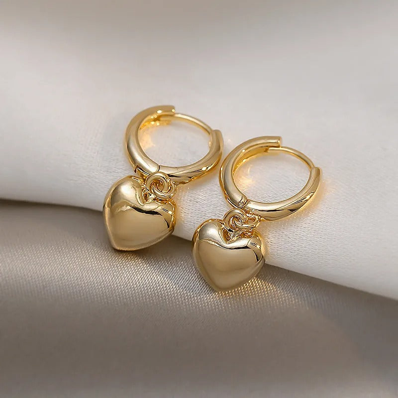 Stylish gold hoop earrings with shiny heart pendants, modern minimalist jewellery displayed on satin fabric background
