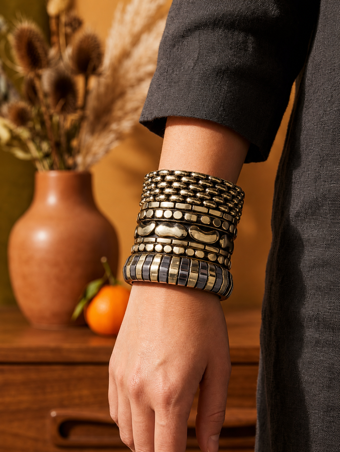 Wrist wearing stacked gold textured bangles against a dark linen sleeve with terracotta vase in background