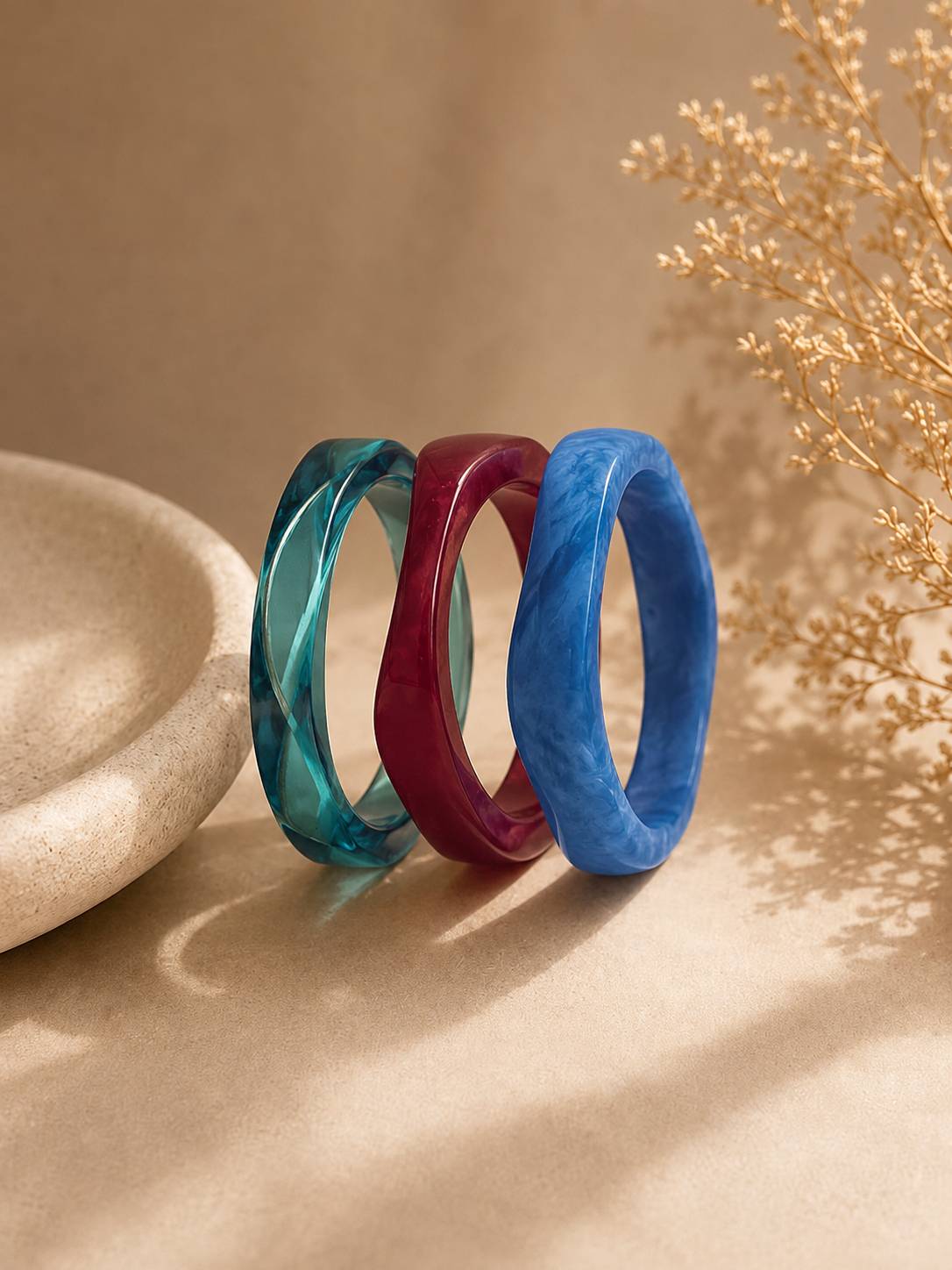 Teal, burgundy and blue marble resin bangles standing upright on a beige surface with dried botanicals