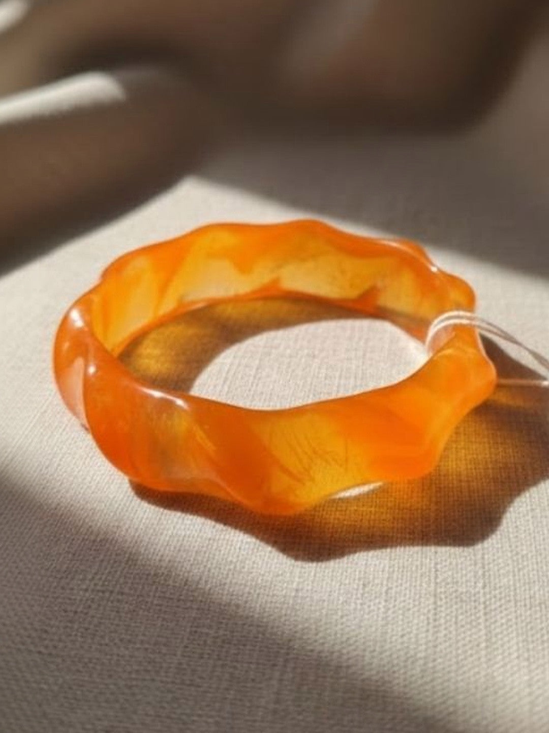 Single orange resin bangle with wavy profile lying flat on a linen fabric surface in natural light