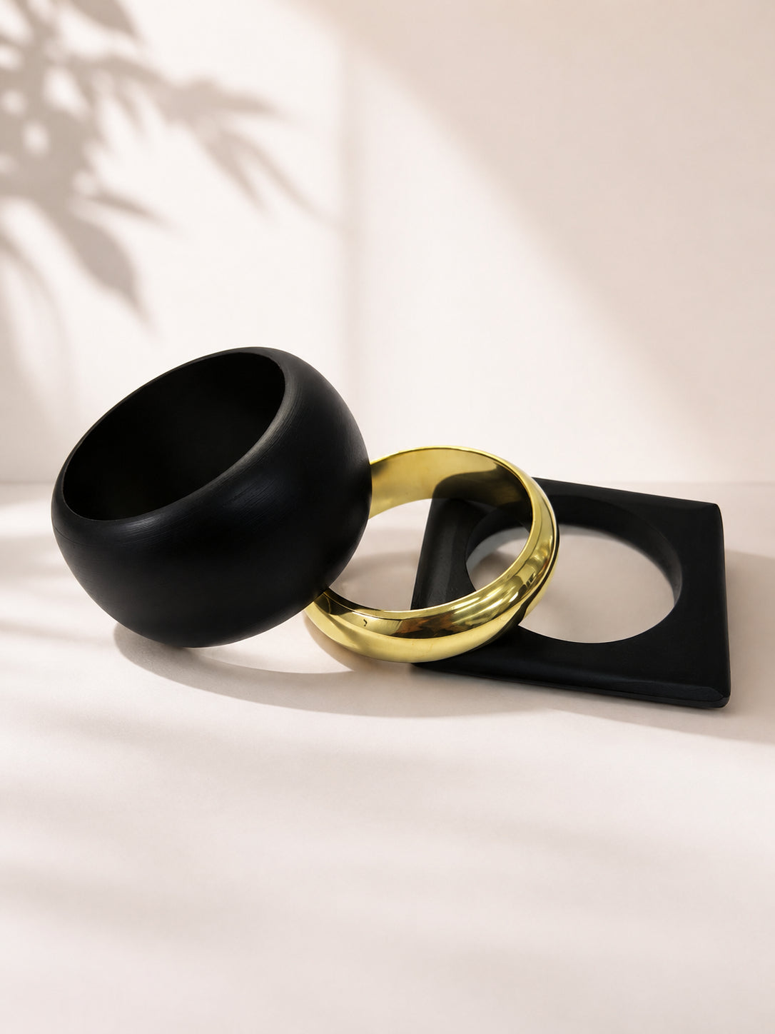 Black dome bangle leaning against gold-tone and square black cuff bangles on a soft beige surface with leaf shadow