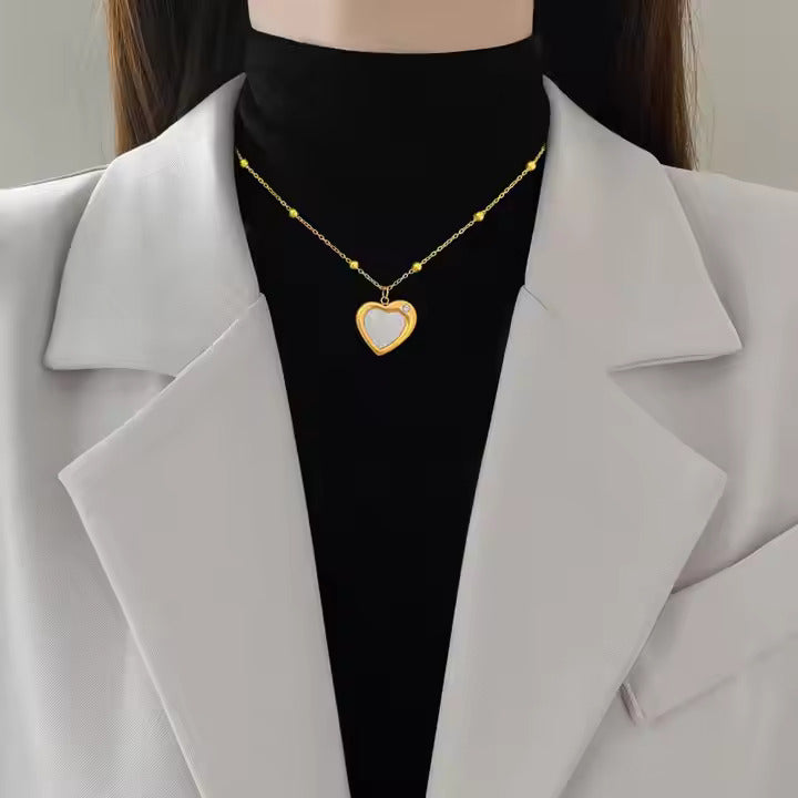Woman wearing a gold beaded chain necklace with a white heart-shaped enamel pendant and diamond accent, styled with a black top and white blazer.