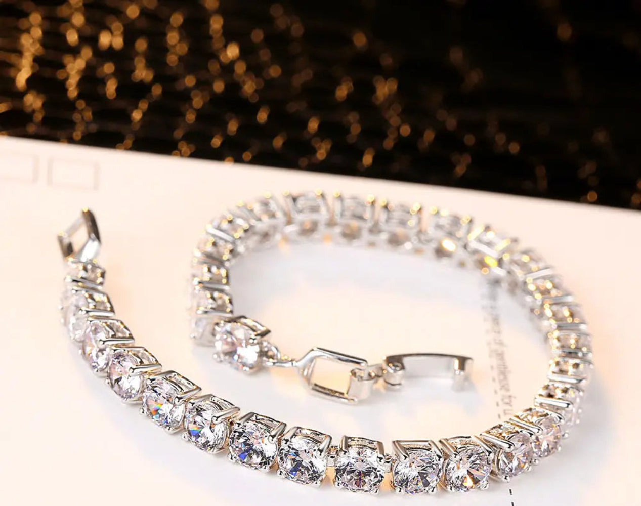 Luxury silver tennis bracelet with sparkling round-cut stones placed on a white background with golden reflections.