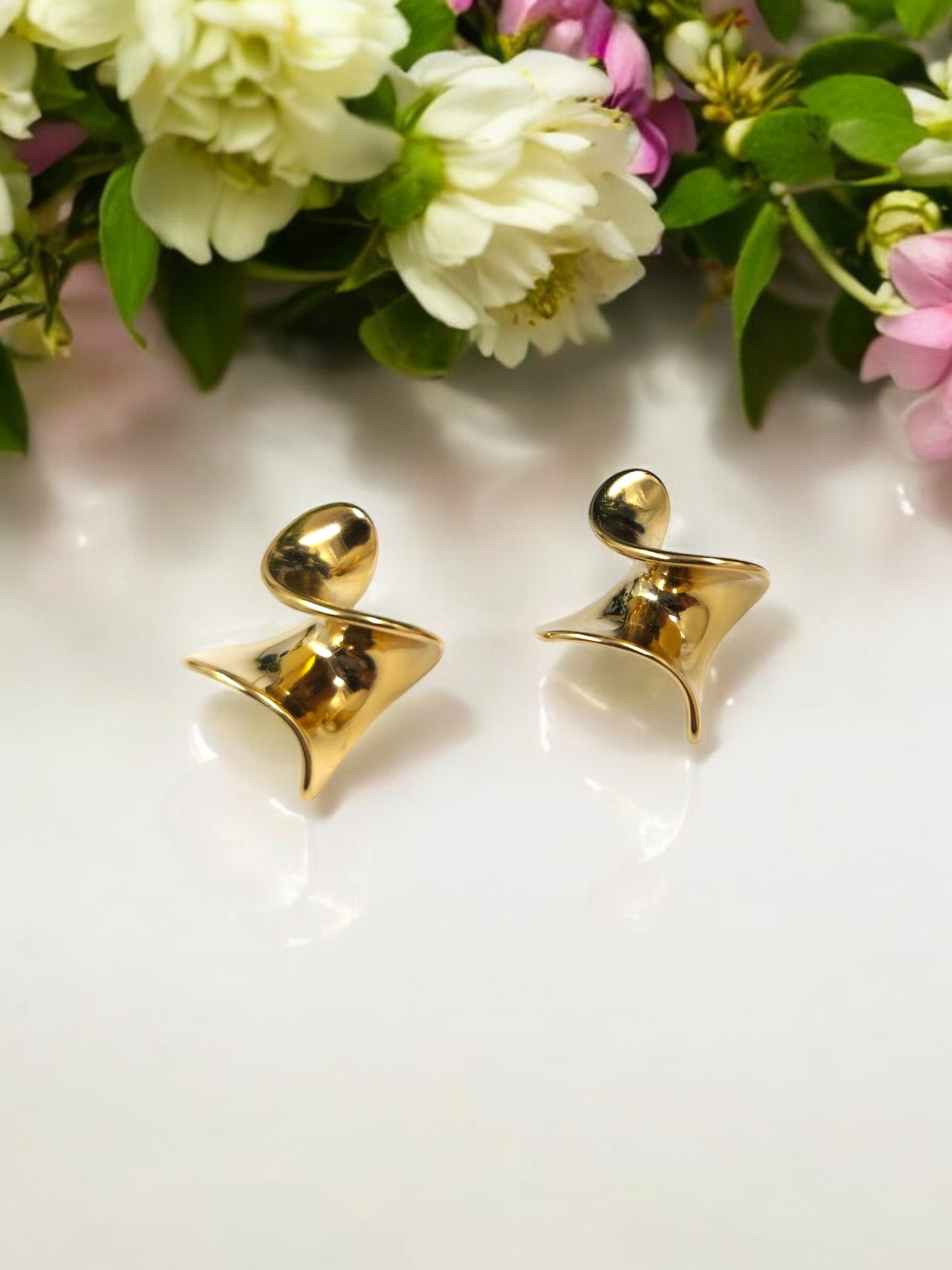 A pair of golden drape earrings with an abstract design, placed on a white surface with flowers in the background.