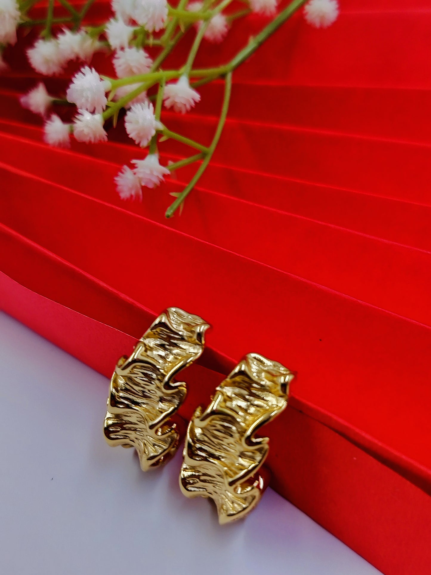 Gold textured leaf-inspired hoop earrings placed on white base with red draped background and baby’s breath flowers.