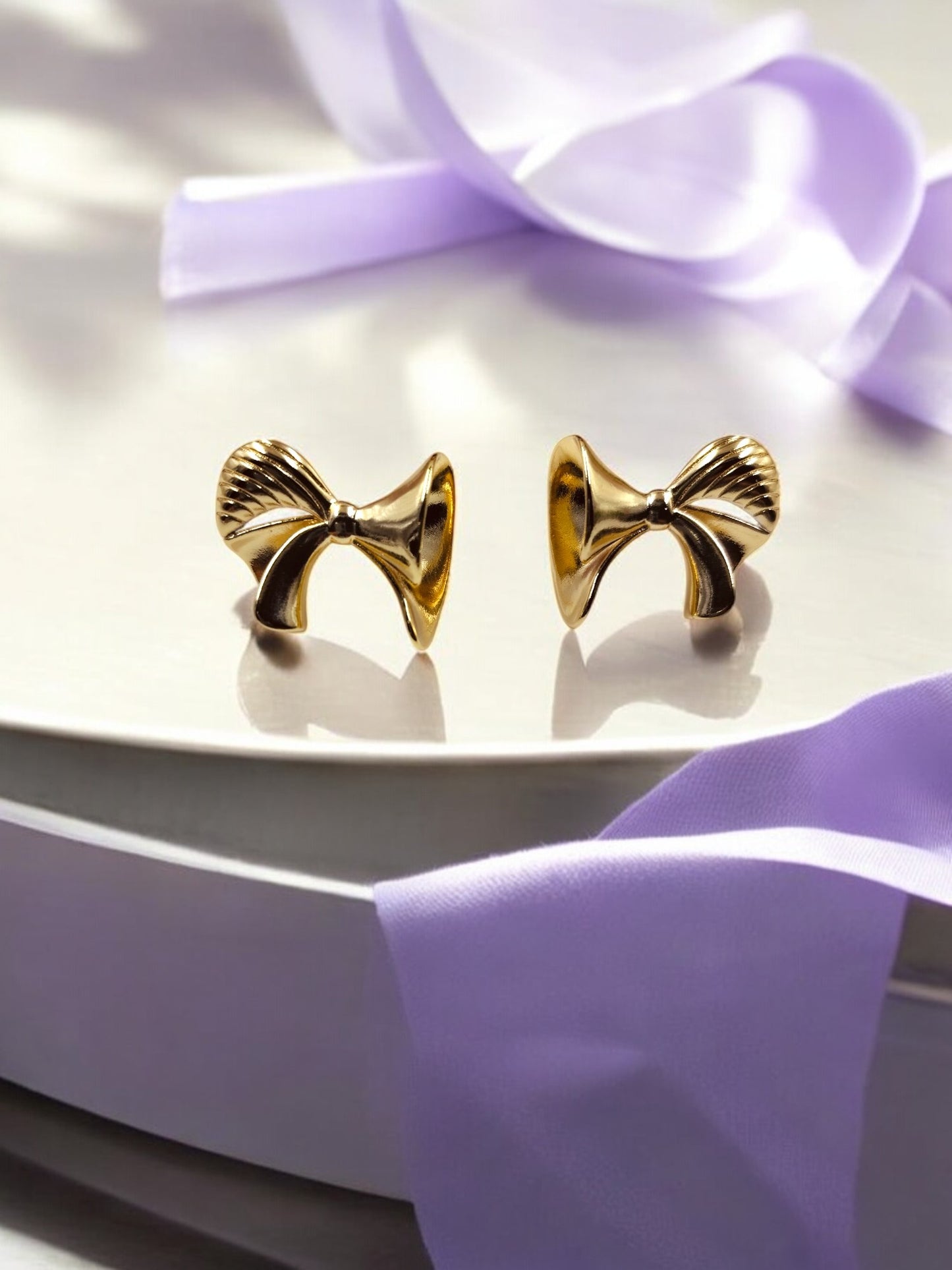 Elegant gold bow-shaped stud earrings with textured detailing, placed on glossy surface with lilac satin ribbon.