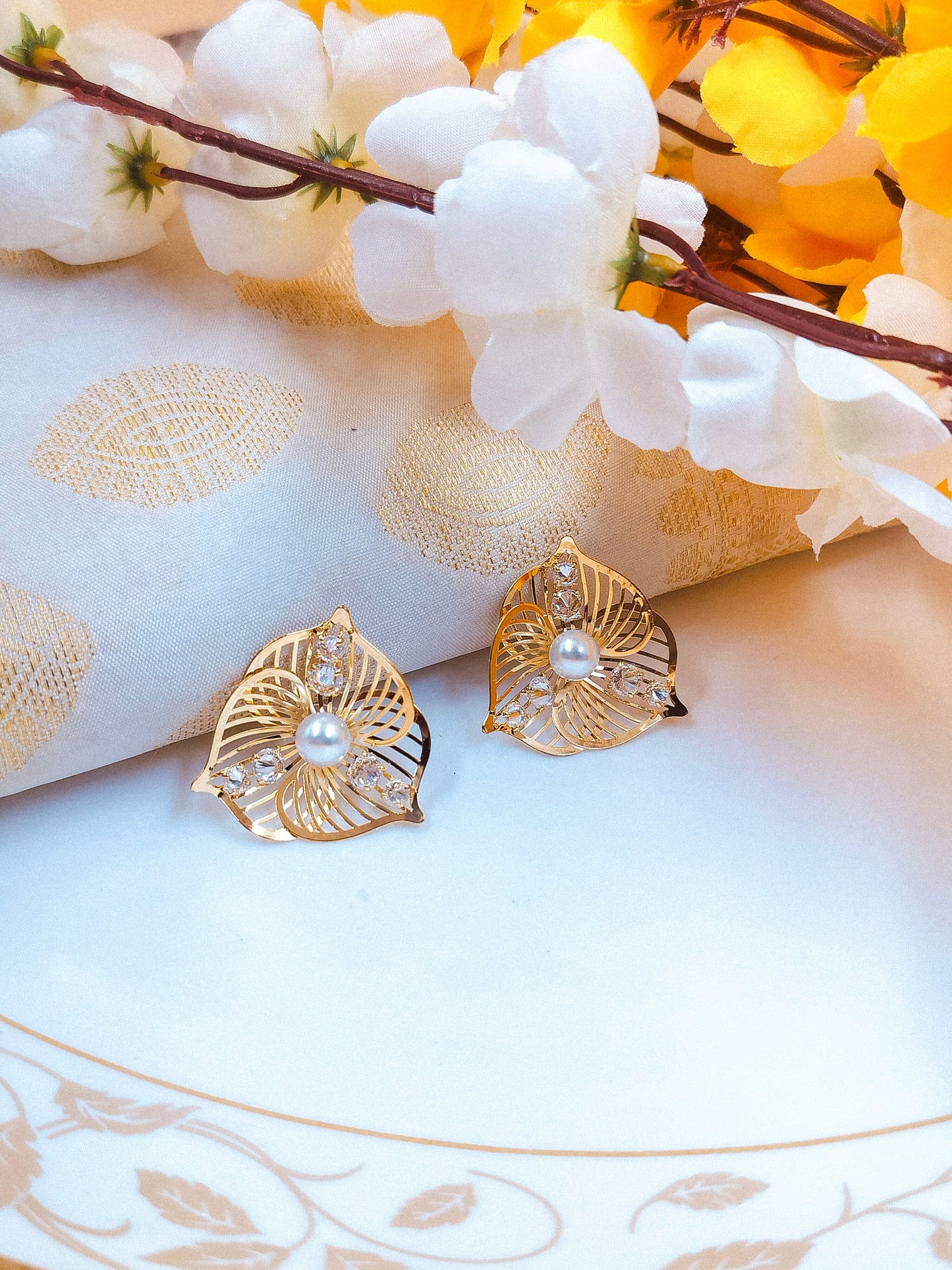 Elegant gold leaf-shaped earrings with pearl center and crystal accents, placed on white fabric with floral decoration.