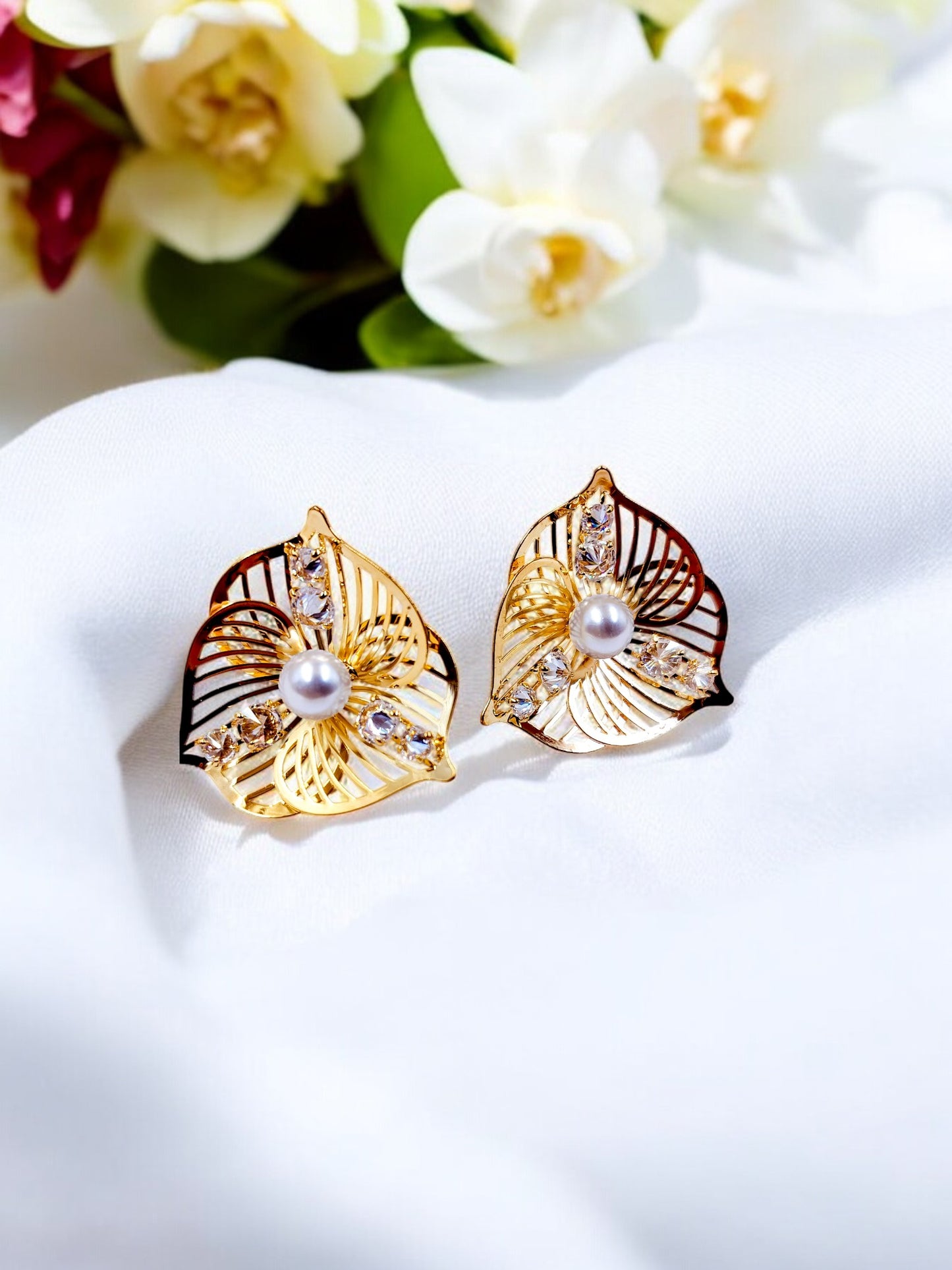 Stylish gold leaf earrings with pearl center and crystal details, displayed on soft white fabric with flowers in background.