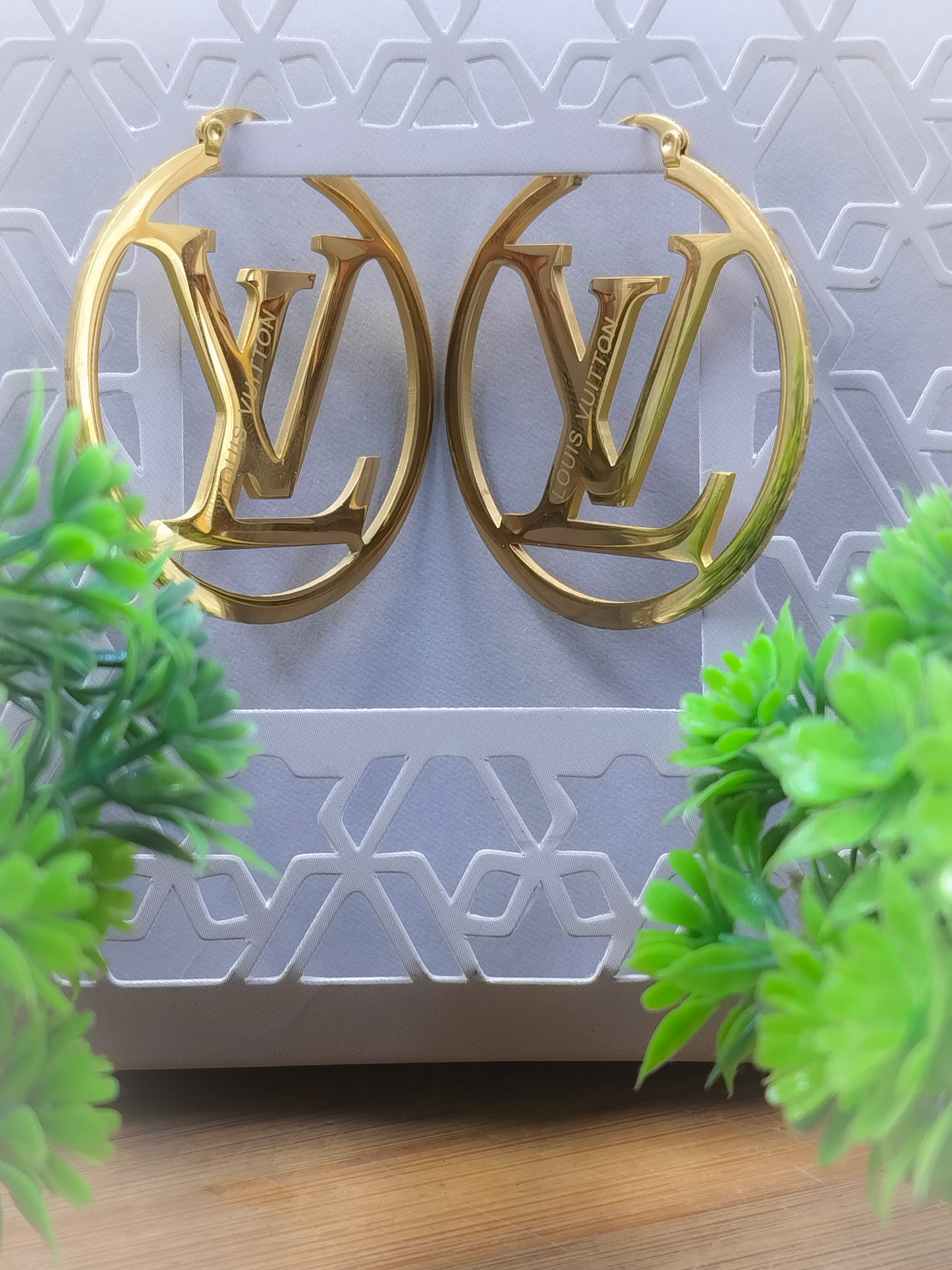Golden LV hoop earrings displayed on textured grey background with green leaves in foreground, luxury designer jewelry.