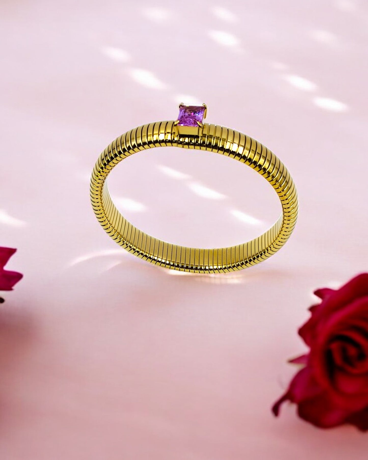 A gold-colored stretchable snake coil elastic bangle bracelet with a single purple stone on a pink background with a rose.