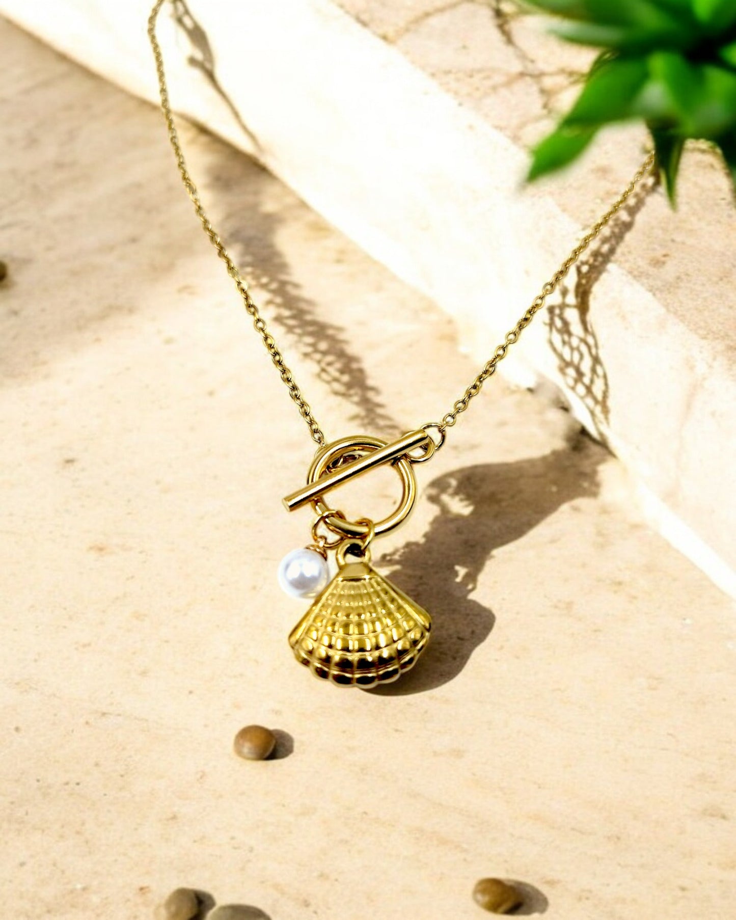 A gold-colored floral pendant necklace with a pearl center displayed on a stone surface with some leaves in the background.