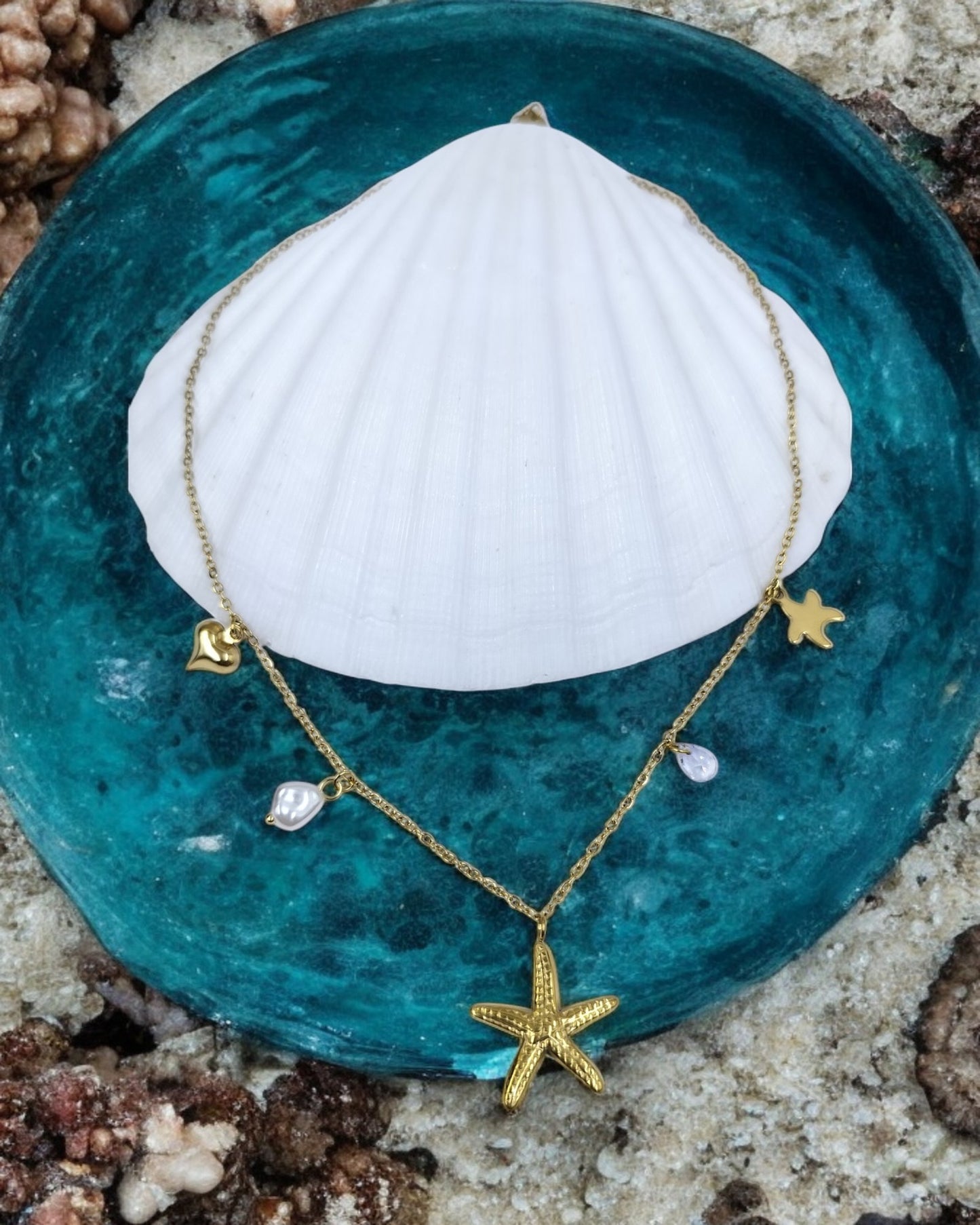 A gold starfish pendant necklace with small oceanic charms and pearl details, displayed on a blue dish with a white shell in the background.