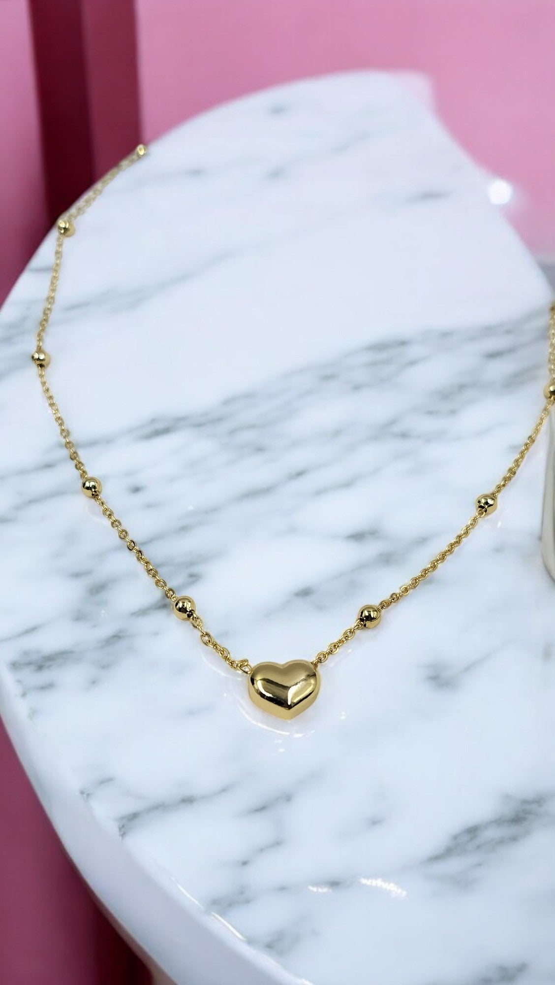 A delicate gold chain necklace with a small heart-shaped pendant, displayed on a marble surface.