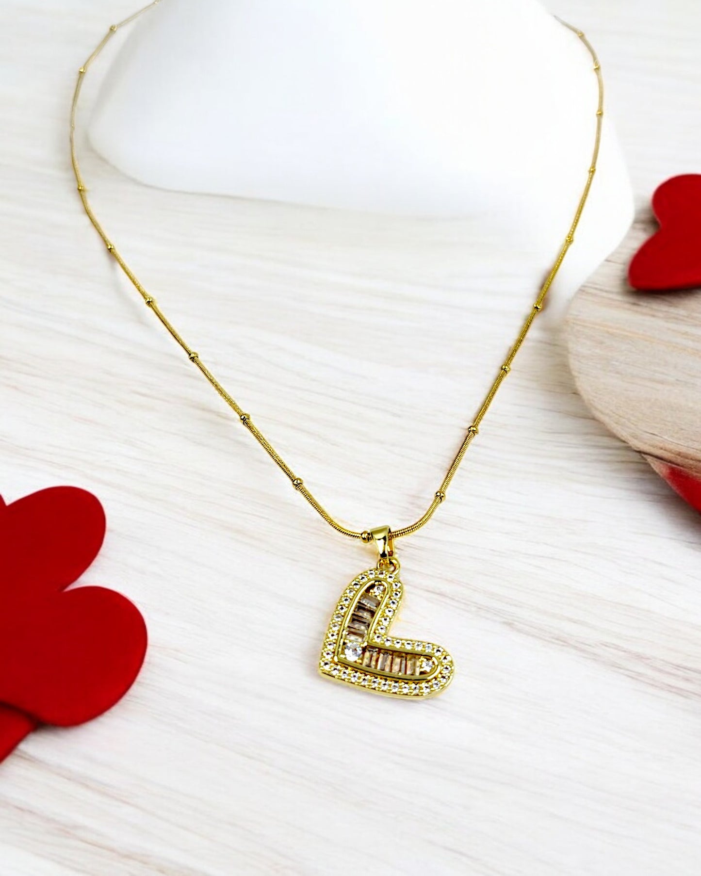 A gold chain necklace with a dainty heart-shaped pendant, displayed on a white surface with red heart-shaped decorations around it.