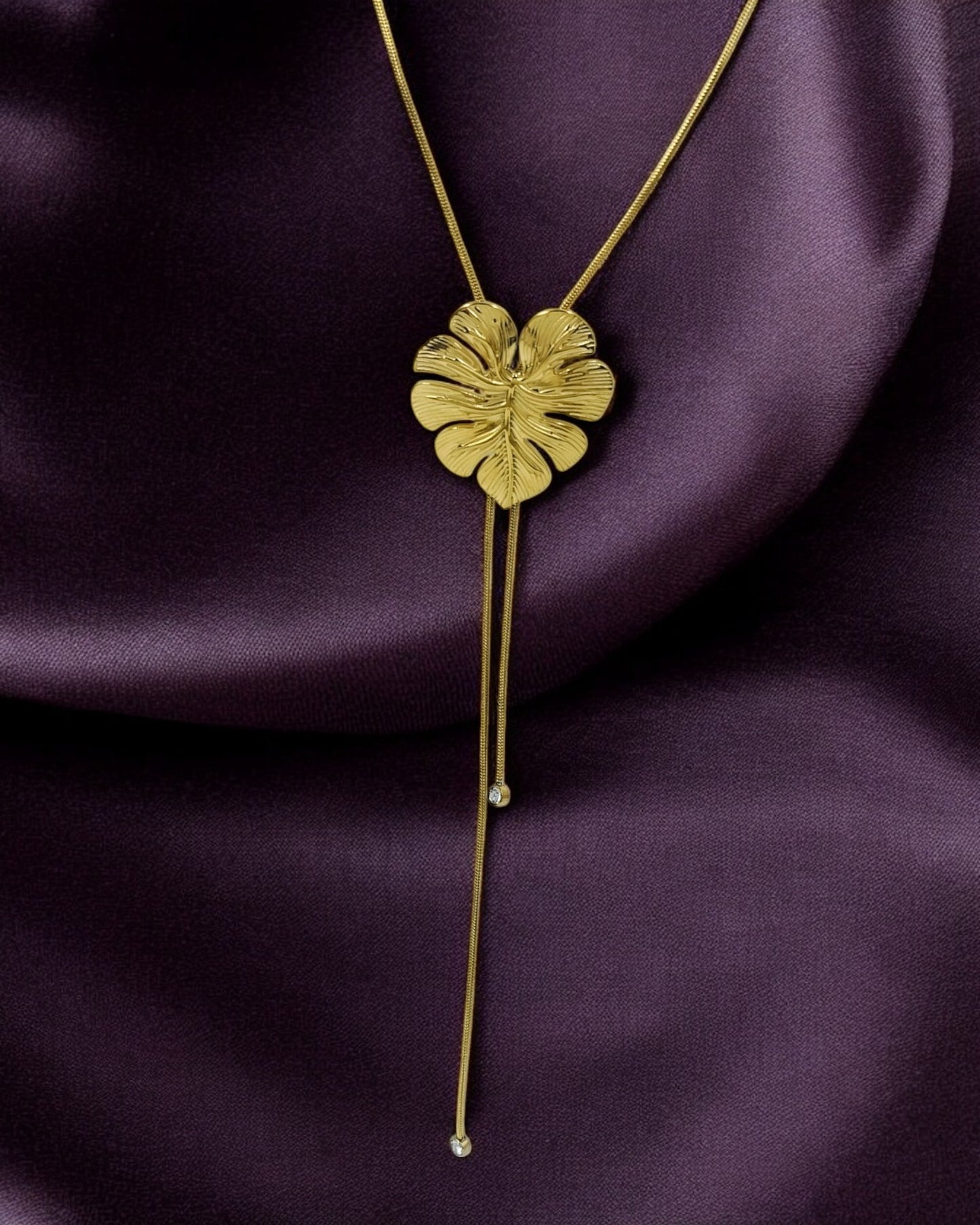 A gold-colored palm leaf pendant on a long Y chain necklace, displayed on a purple background.