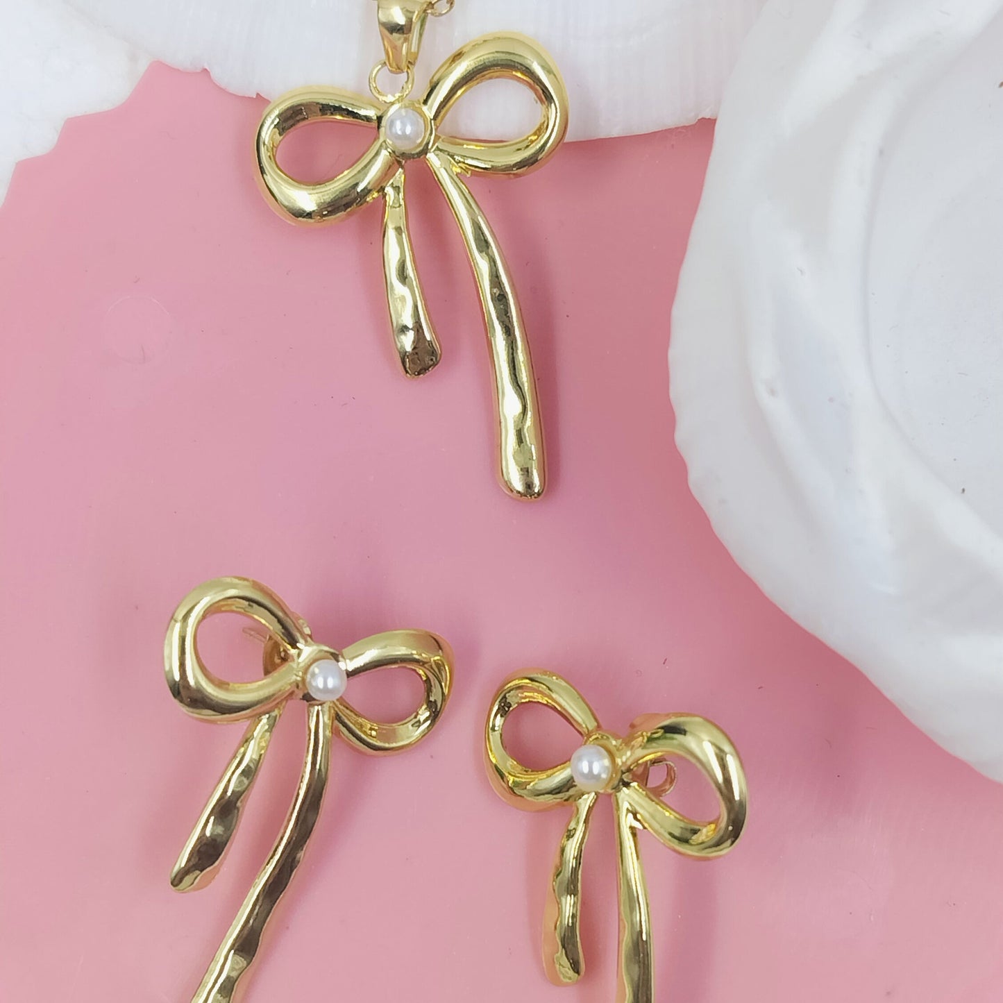 A set of gold-colored bow knot pendant jewelry, including a necklace and earrings, displayed on a pink surface.