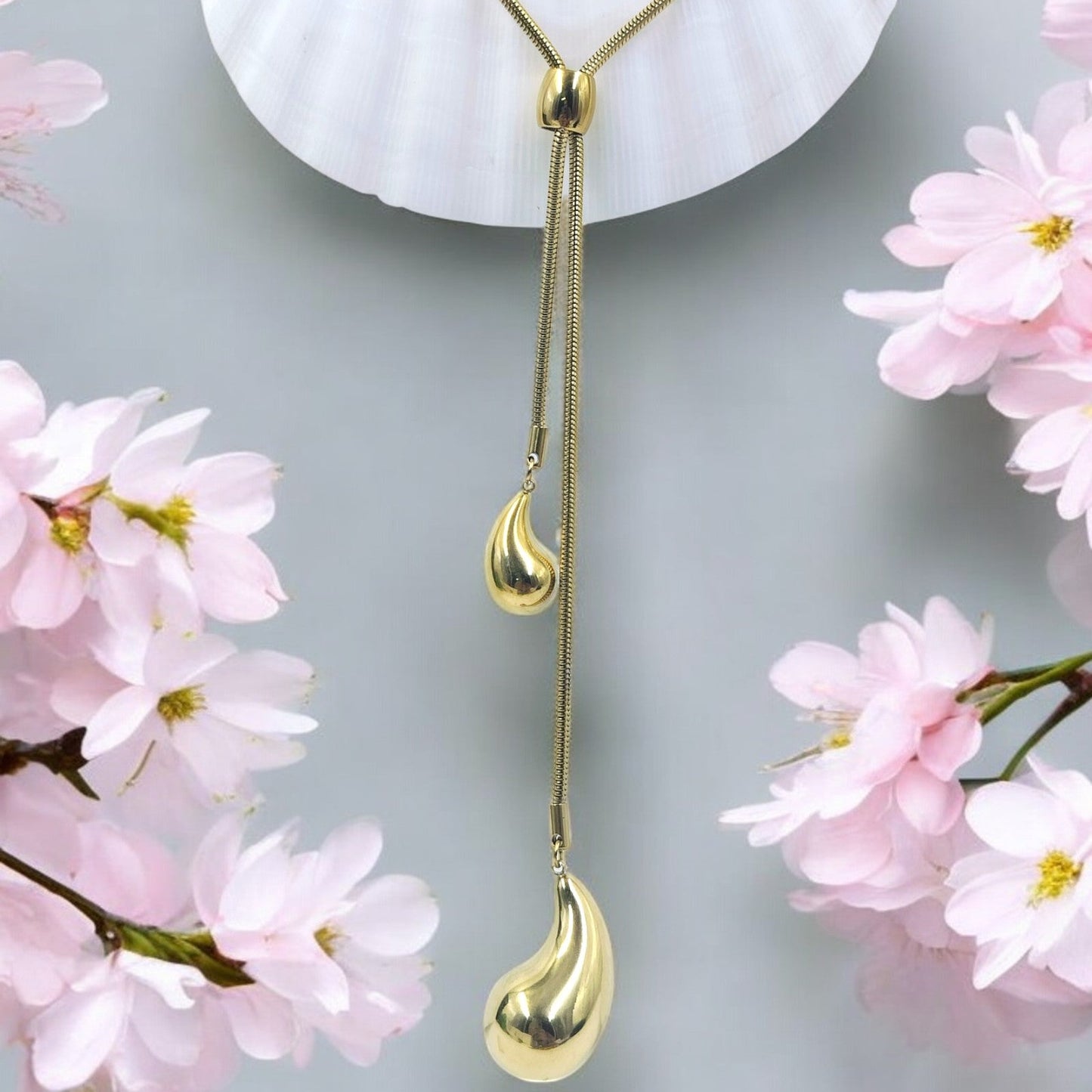 A gold-colored long Y chain necklace with a droplet pendant, displayed against a background with pink floral elements.