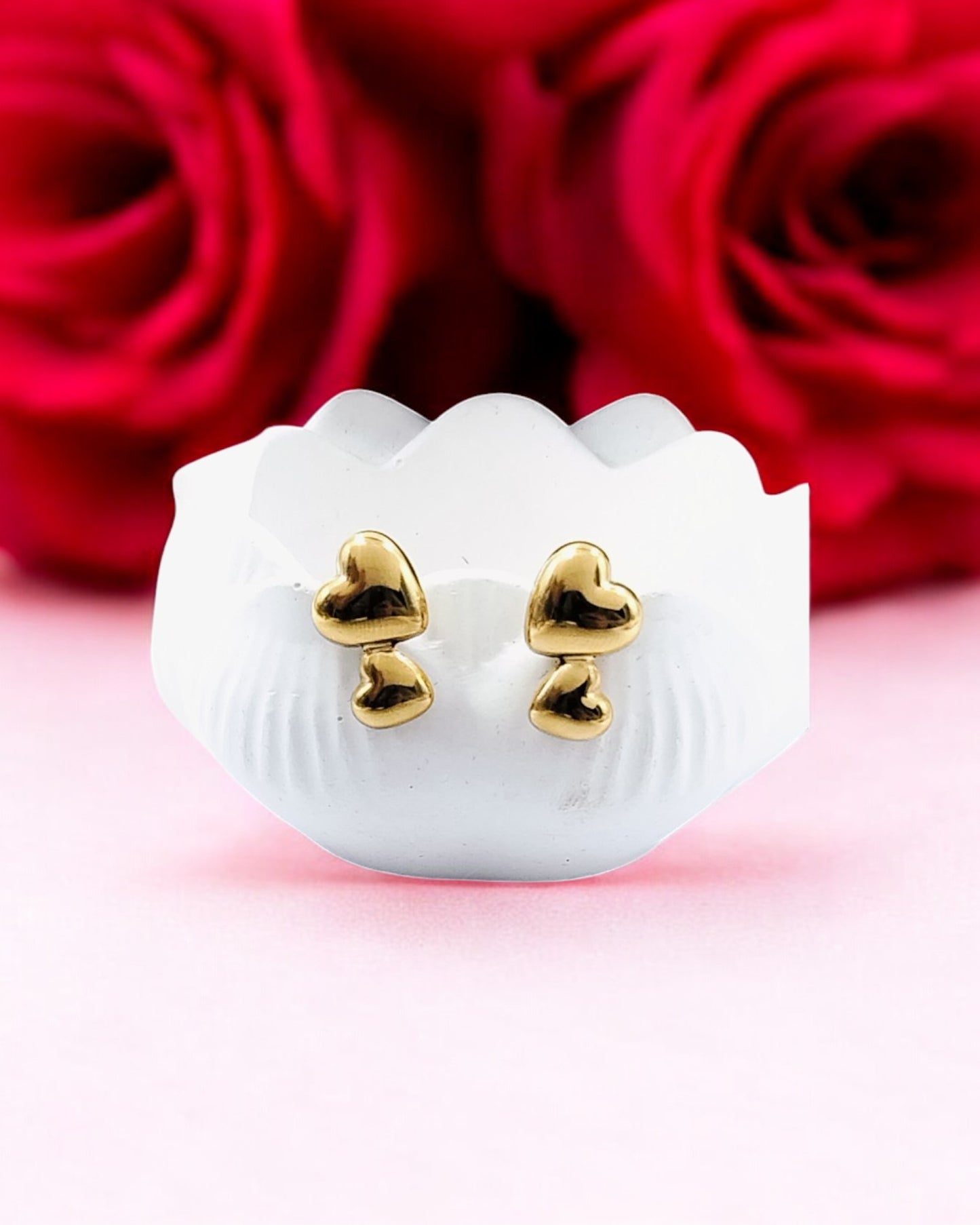 A pair of gold-colored double heart stud earrings displayed in front of a red rose.