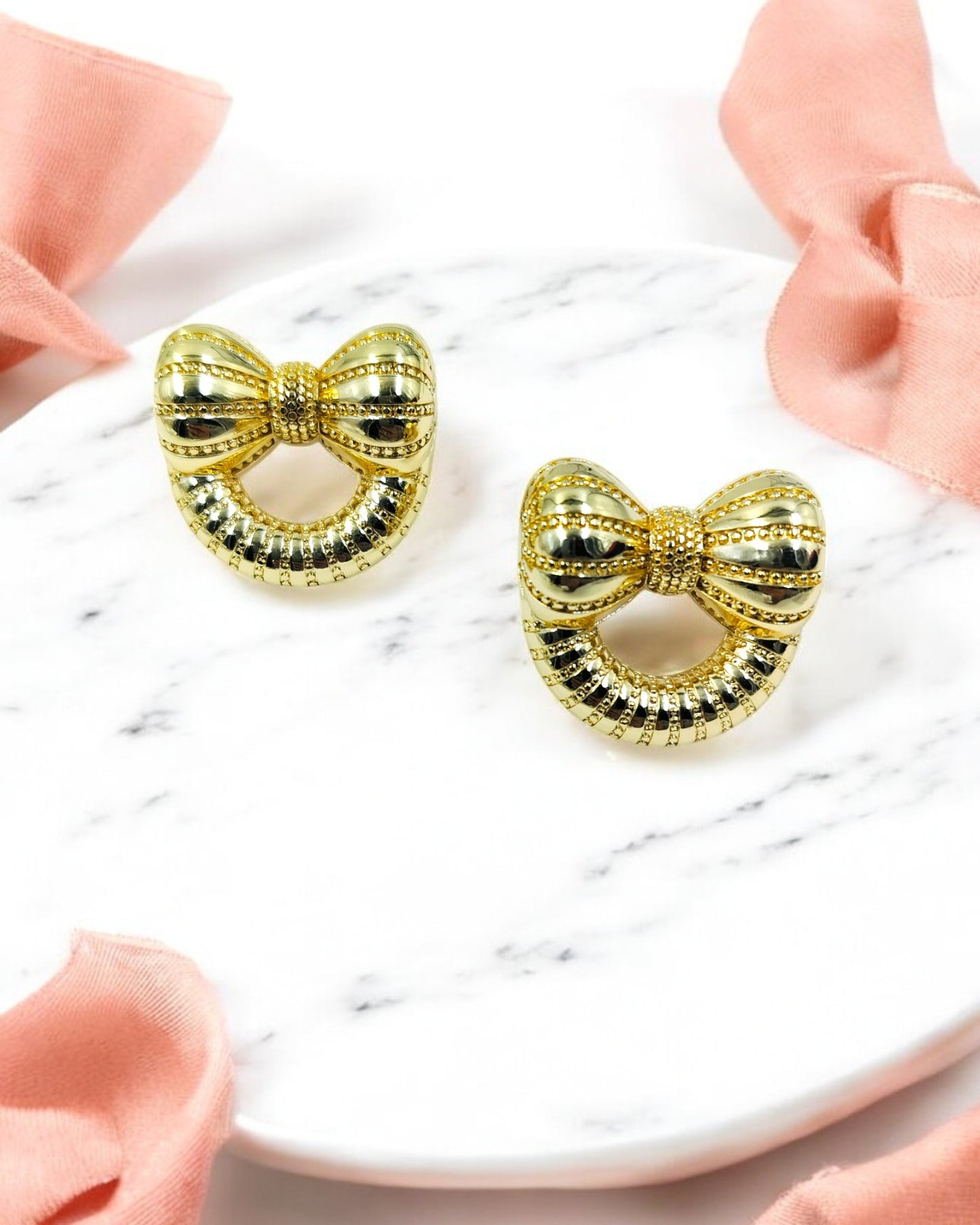 A pair of gold-colored bow-shaped earrings on a white surface with a pink ribbon beside them.