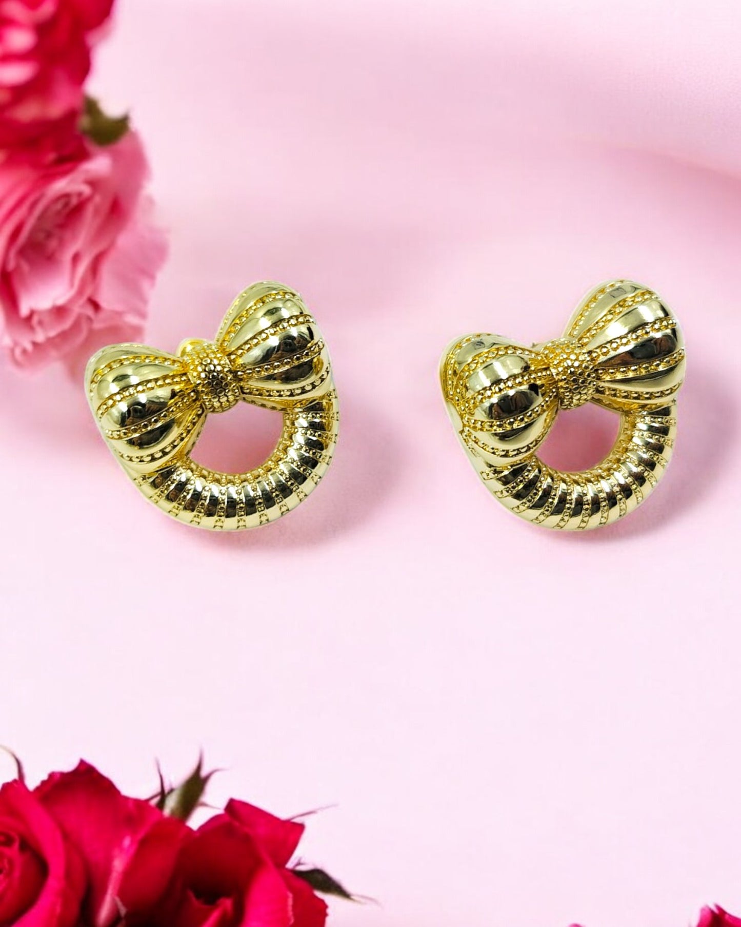 Golden bow knot earrings with textured circular design on pink background with roses.