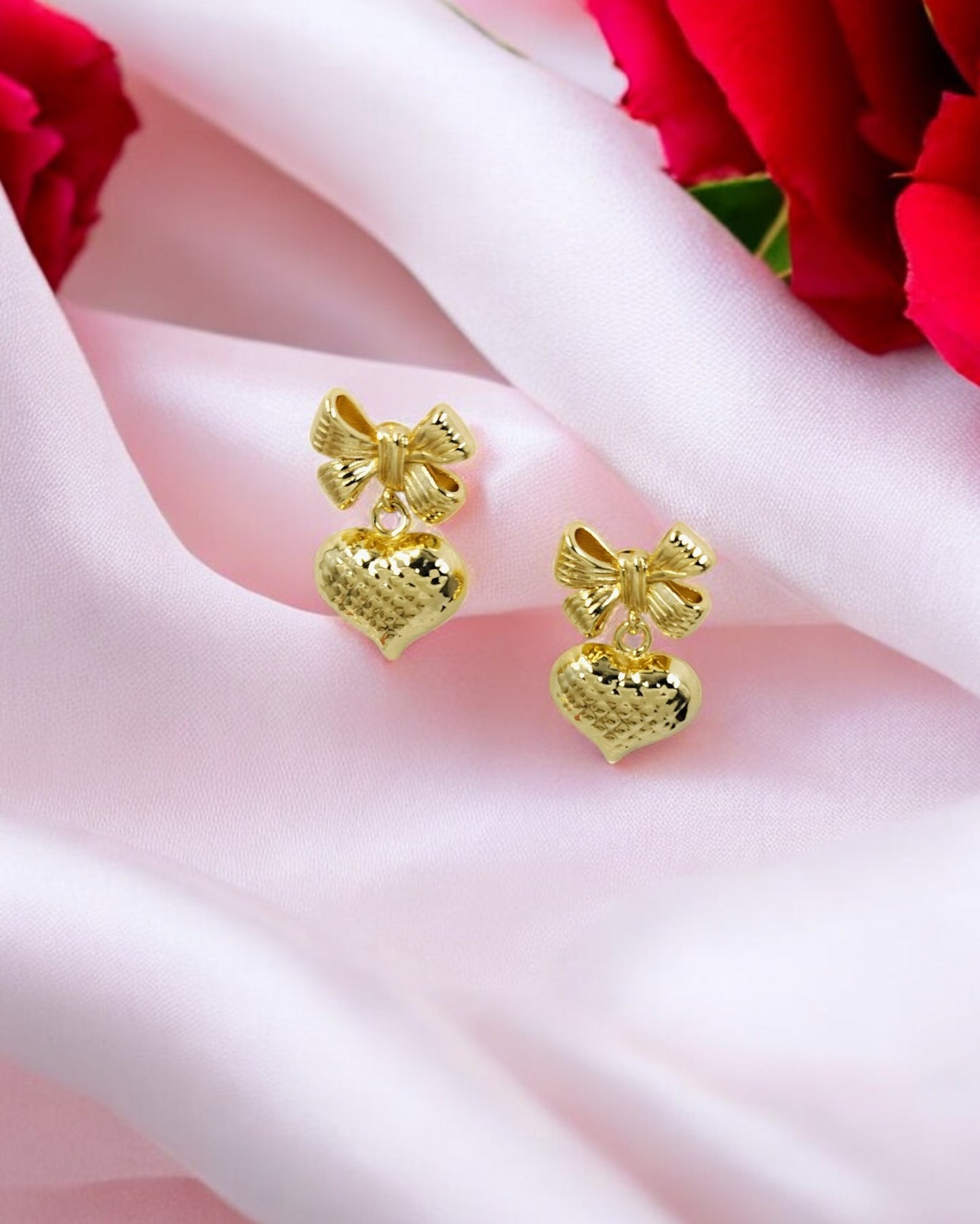 Elegant gold bow and heart earrings on pink silk with roses, romantic jewelry gift for women.