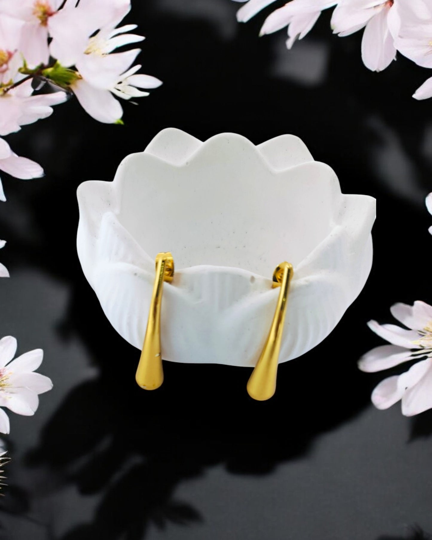 Modern gold-plated earrings with a curved, polished drop shape, sitting in a white ceramic bowl on a black surface with flowers.