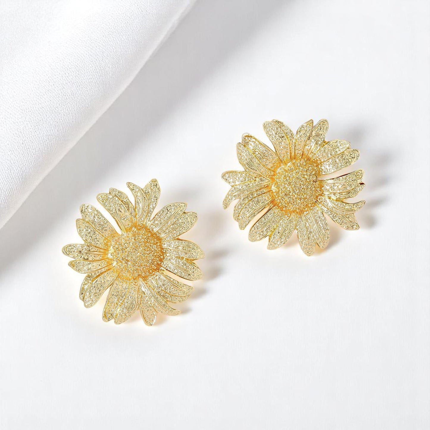 Golden sunflower-shaped earrings displayed on a plain white background with a folded cloth beside them.
