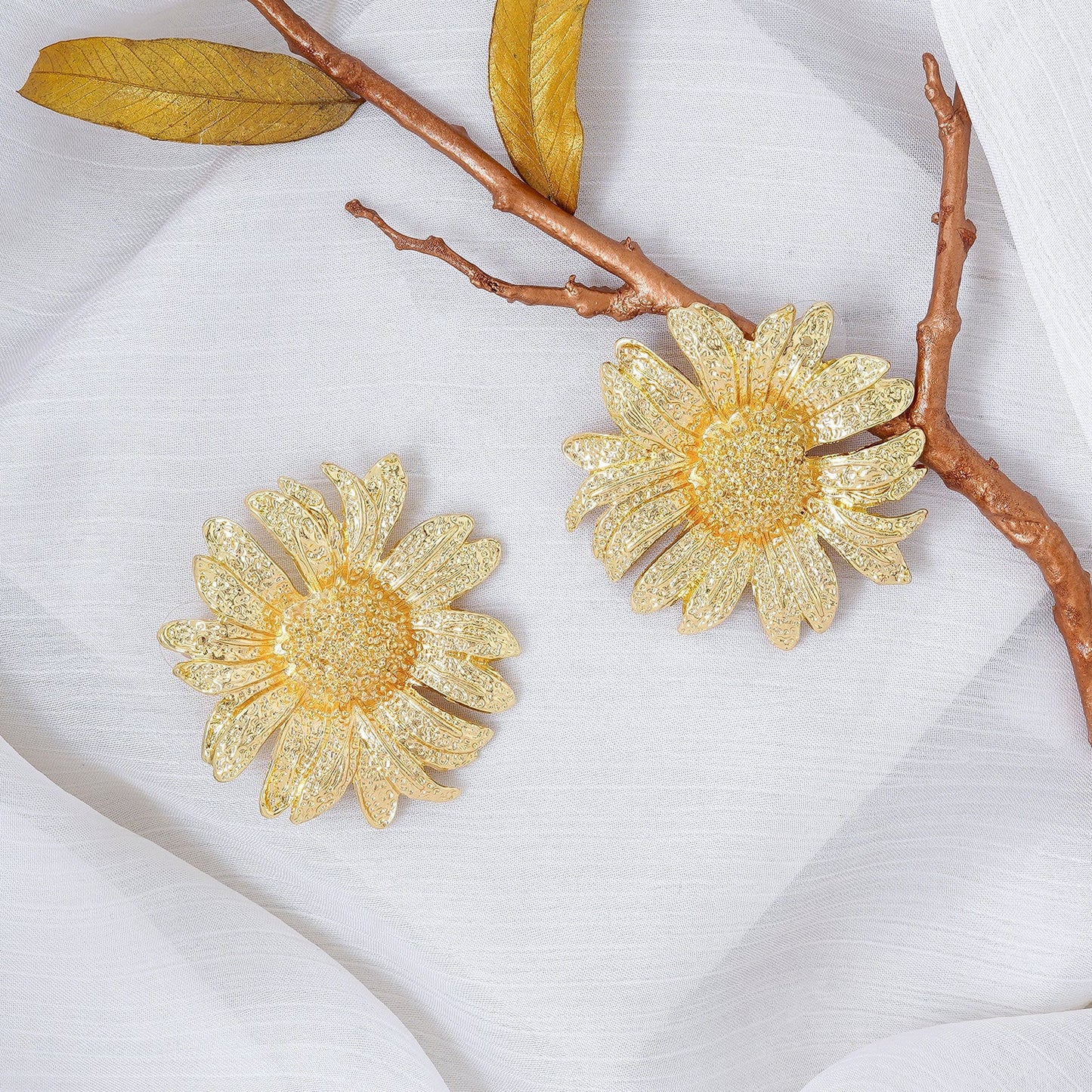 Stunning gold floral statement earrings placed on a soft white textured fabric with golden leaves and a twig for an elegant and artistic display.