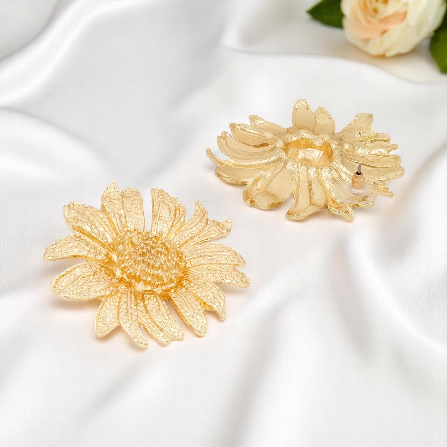 Pair of golden sunflower-shaped earrings placed on soft white satin fabric with a rose in the background.