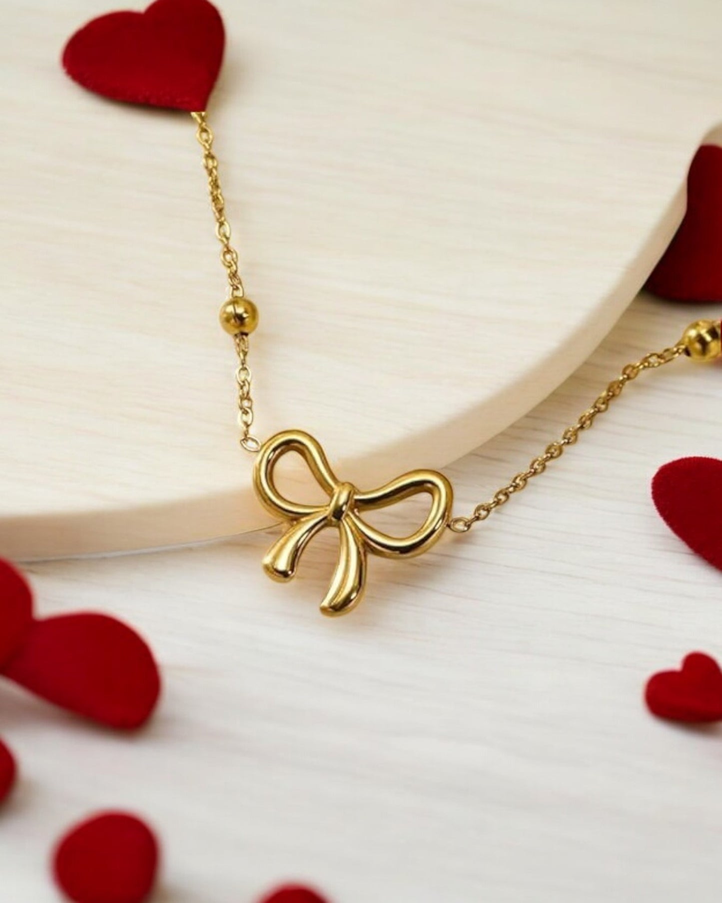 A delicate gold necklace with a bow pendant displayed on a white surface with scattered red hearts.