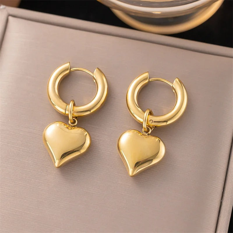 Elegant gold hoop earrings with dangling heart charms, shiny minimalist jewellery displayed on a soft neutral background