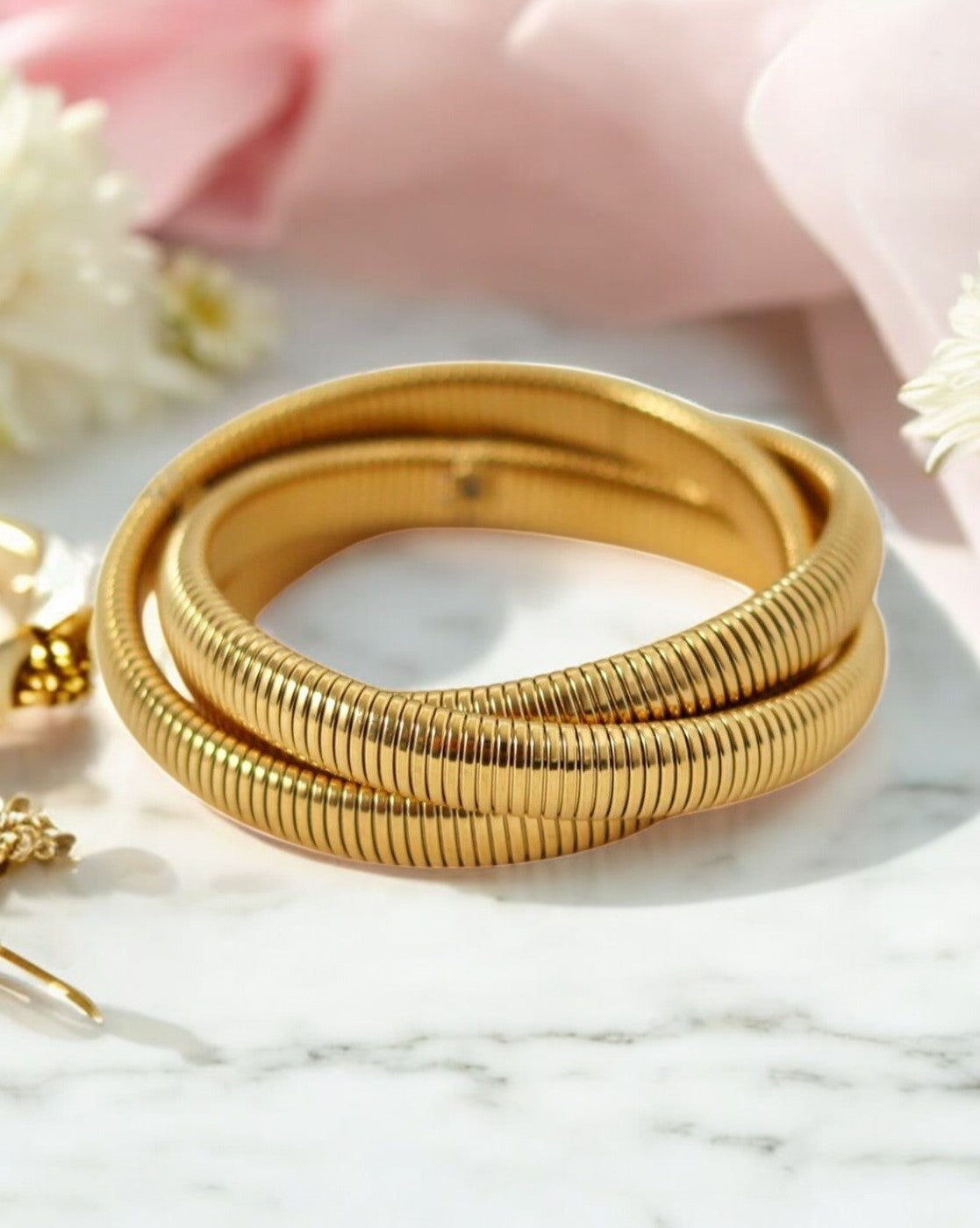 A gold-colored triple layer interlock snake coil elastic bangle bracelet displayed on a marble surface with a pink cloth in the background.