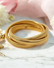 A gold-colored triple layer interlock snake coil elastic bangle bracelet displayed on a marble surface with a pink cloth in the background.