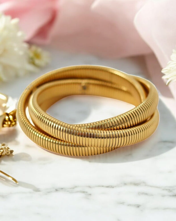 A gold-colored triple layer interlock snake coil elastic bangle bracelet displayed on a marble surface with a pink cloth in the background.