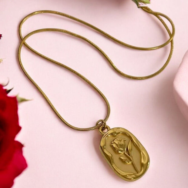 A gold-colored necklace with a heart-shaped pendant featuring a flower design, displayed against a pink background with red roses.