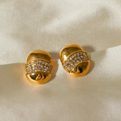 A pair of gold-tone dome-shaped earrings with a wave-like band encrusted with sparkling rhinestones on a white background.