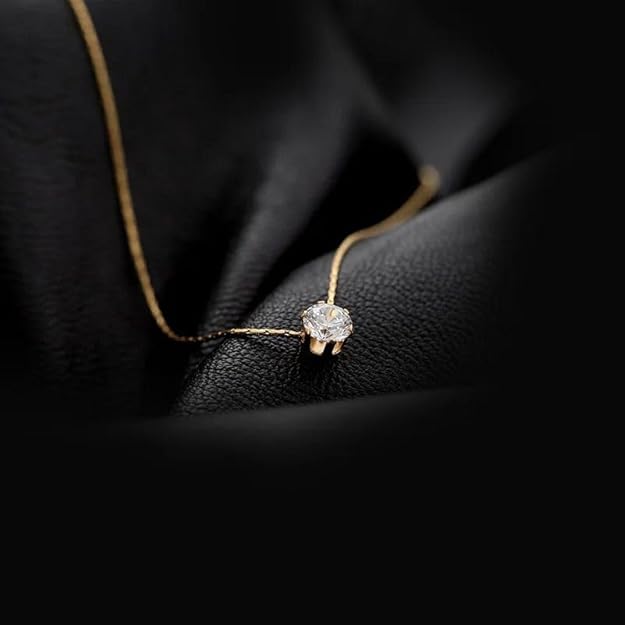 Minimalist gold chain necklace with solitaire diamond pendant placed on black fabric background.