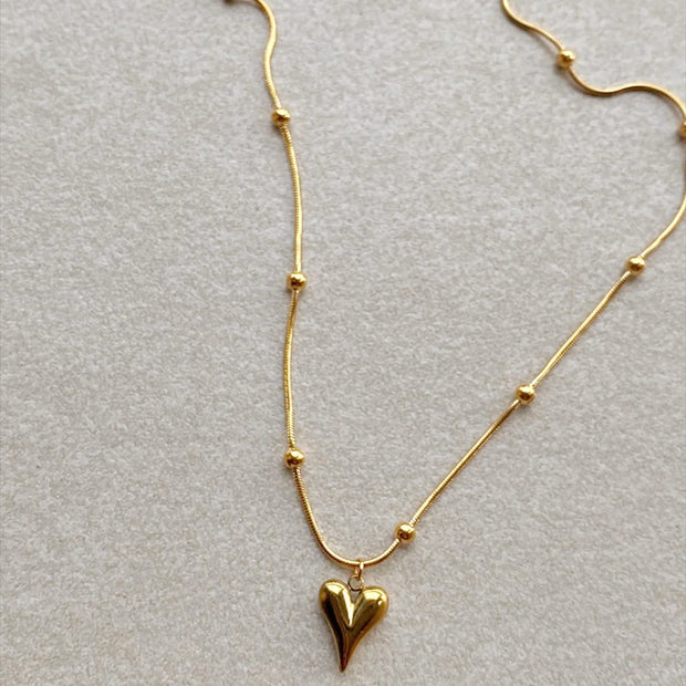 Elegant gold necklace with a central heart pendant and delicate bead accents on fine chain, displayed on soft beige background.