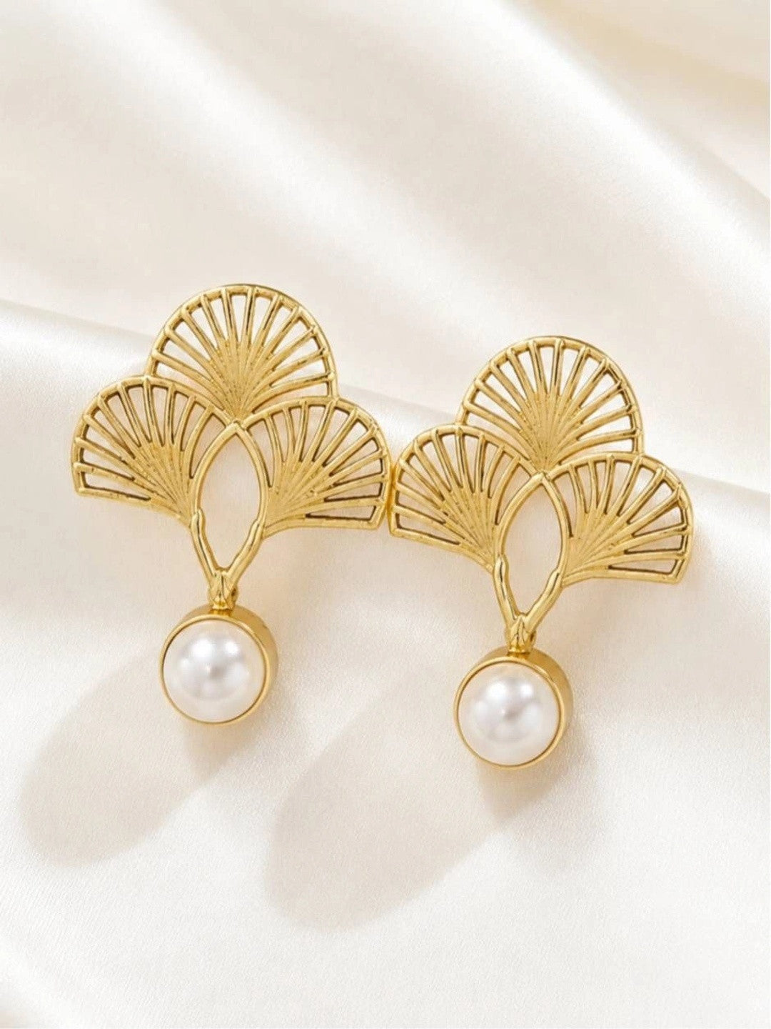 Close-up of two gold openwork fan earrings with pearl drops on white fabric