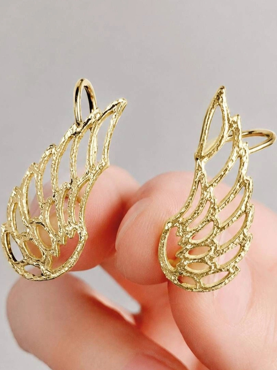 Close-up of two gold wing ear cuffs held between fingers showing textured hammered filigree detail