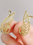 Close-up of two gold wing ear cuffs held between fingers showing textured hammered filigree detail