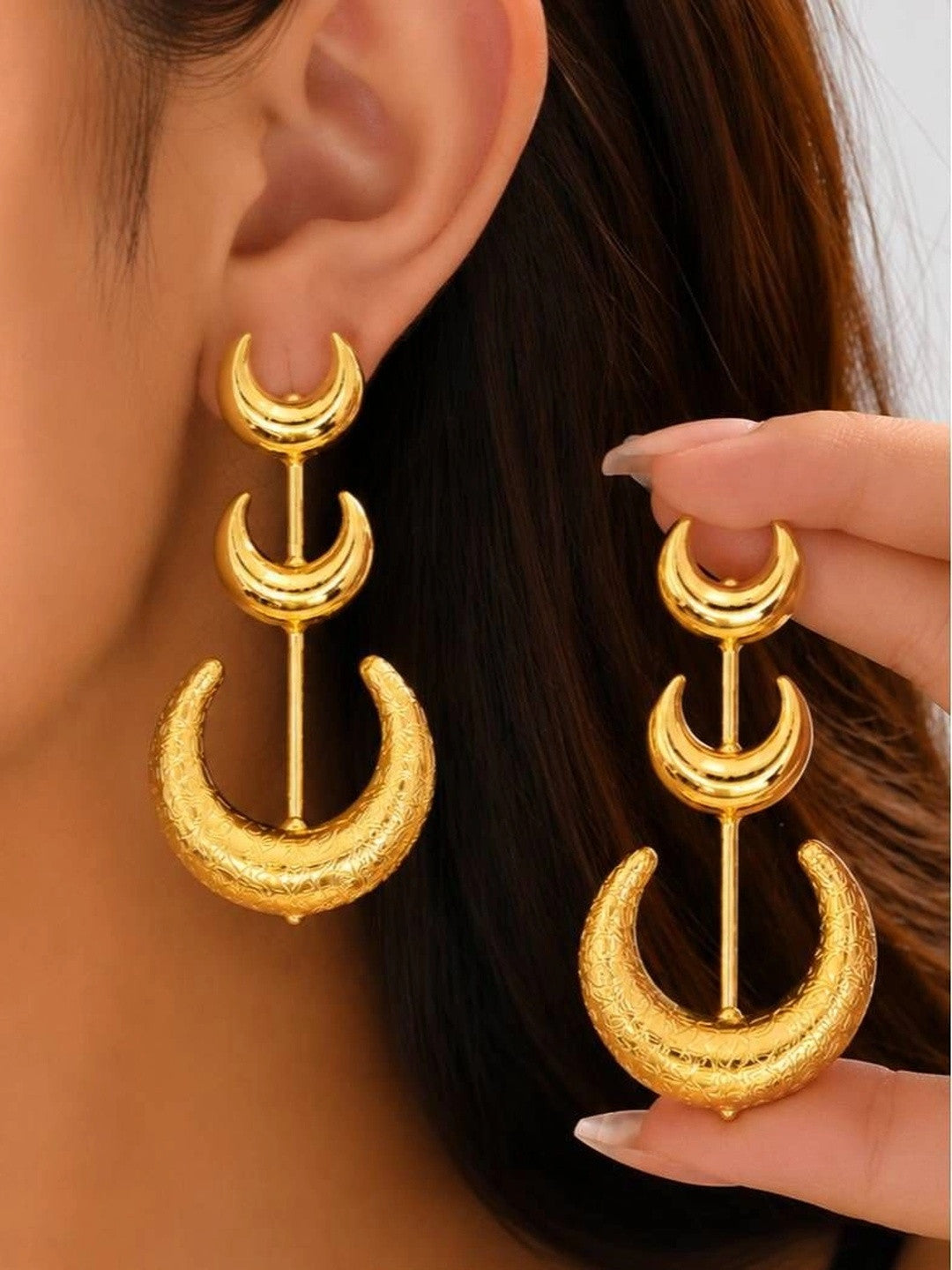Gold triple crescent moon drop earring worn on ear beside a hand holding the second earring
