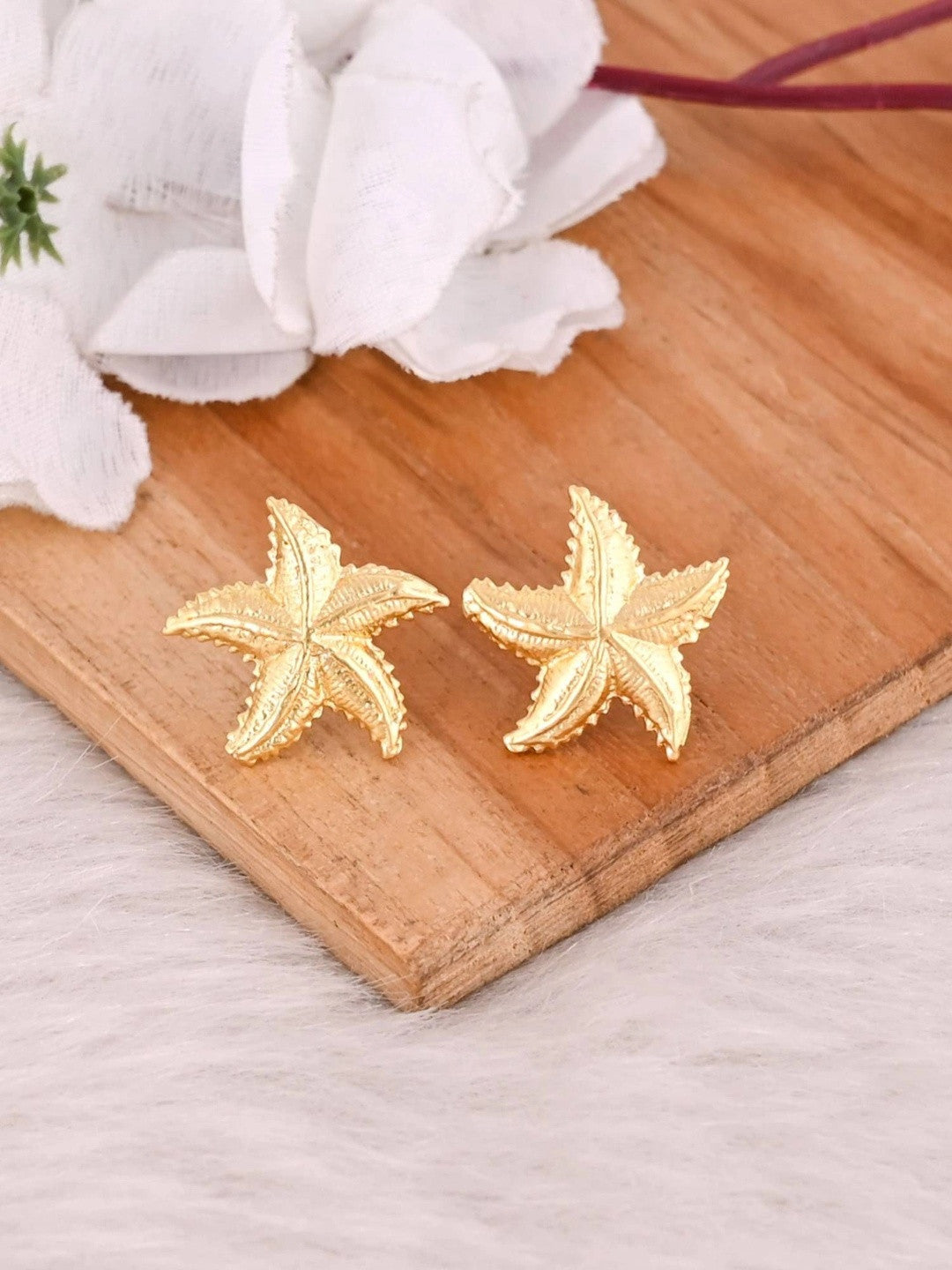Pair of gold-plated starfish stud earrings laid flat on a wooden board, white flower prop in background