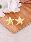 Pair of gold-plated starfish stud earrings laid flat on a wooden board, white flower prop in background