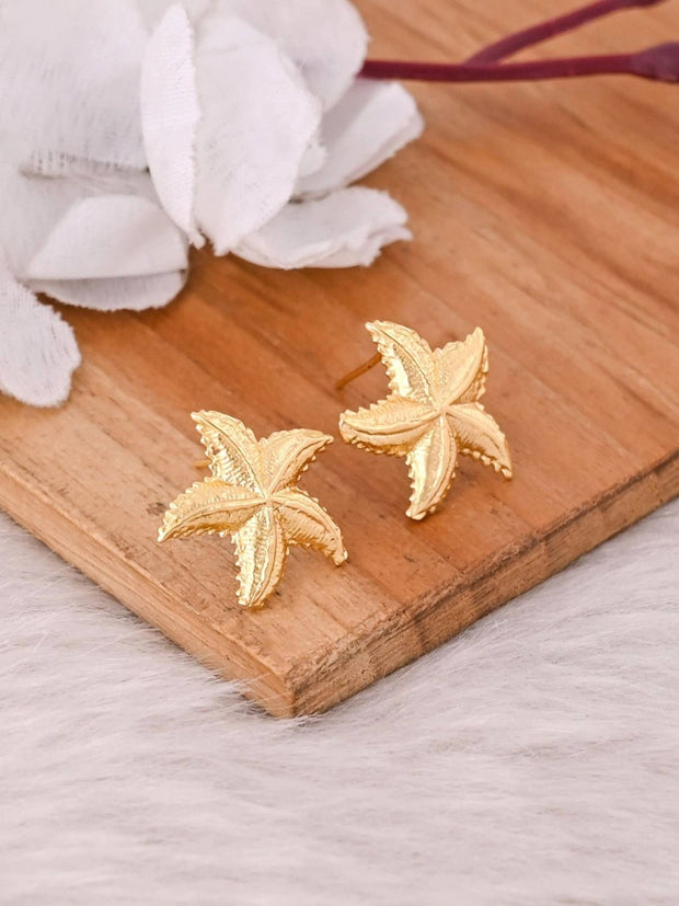 Two gold starfish stud earrings displayed side by side on a wooden surface showing textured arm detail
