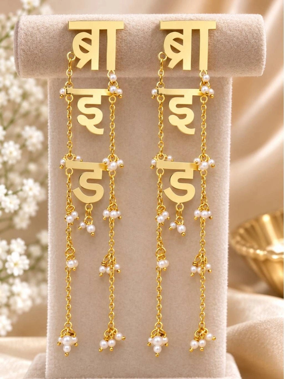 Pair of gold-plated Hindi script bride earrings displayed upright on a beige jewellery stand with pearl bead chain tassels
