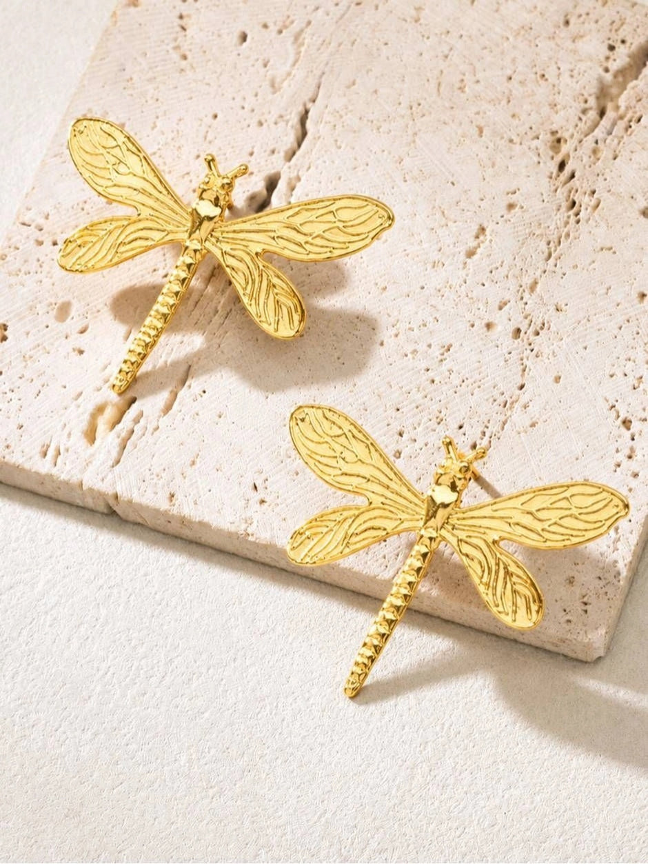 Pair of gold-plated dragonfly stud earrings displayed flat on a beige travertine tile