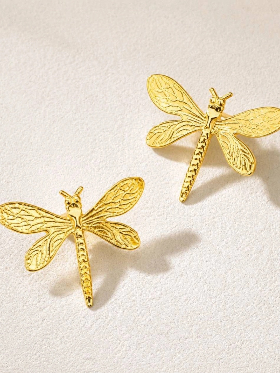 Two gold dragonfly stud earrings shown face-up on a white textured surface highlighting wing detail