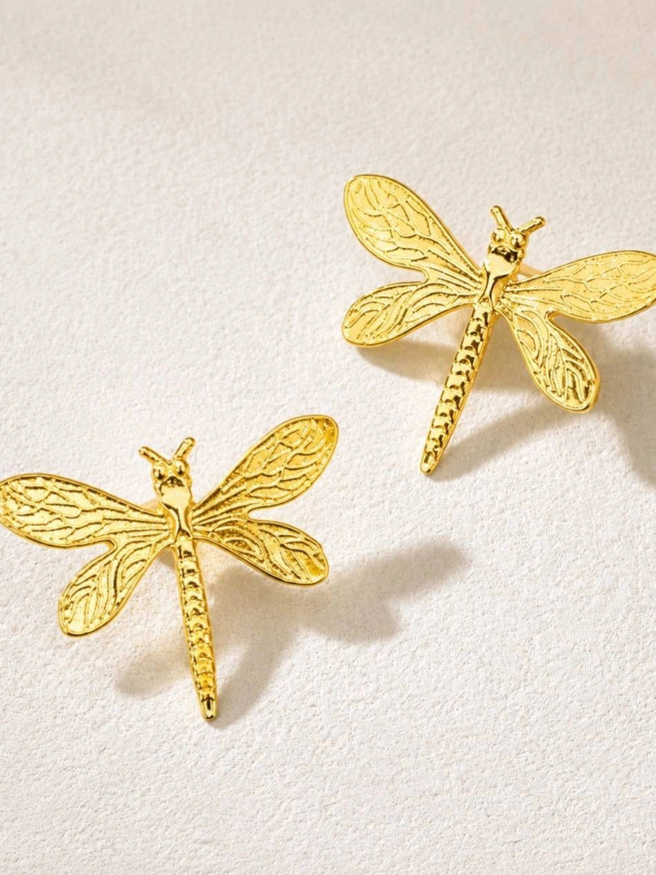 Two gold dragonfly stud earrings shown face-up on a white textured surface highlighting wing detail